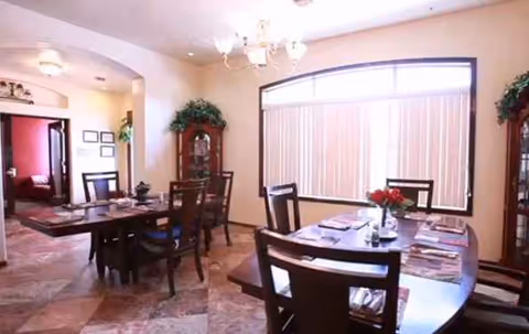 Elegant dining room with wooden tables, chairs, chandeliers, plants, and large window blinds.