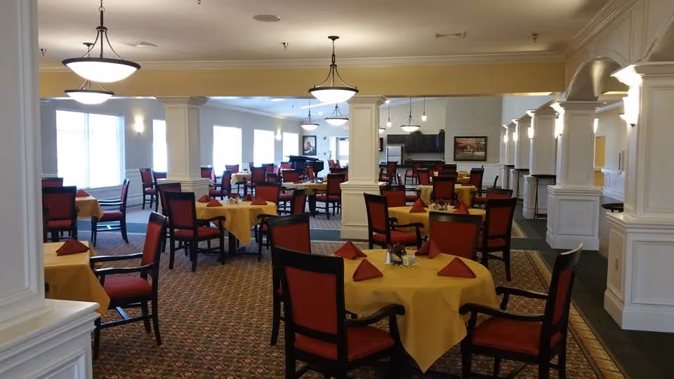 Spacious dining room with round tables covered in yellow tablecloths, red-upholstered chairs, folded red napkins, and white columns under hanging light fixtures.