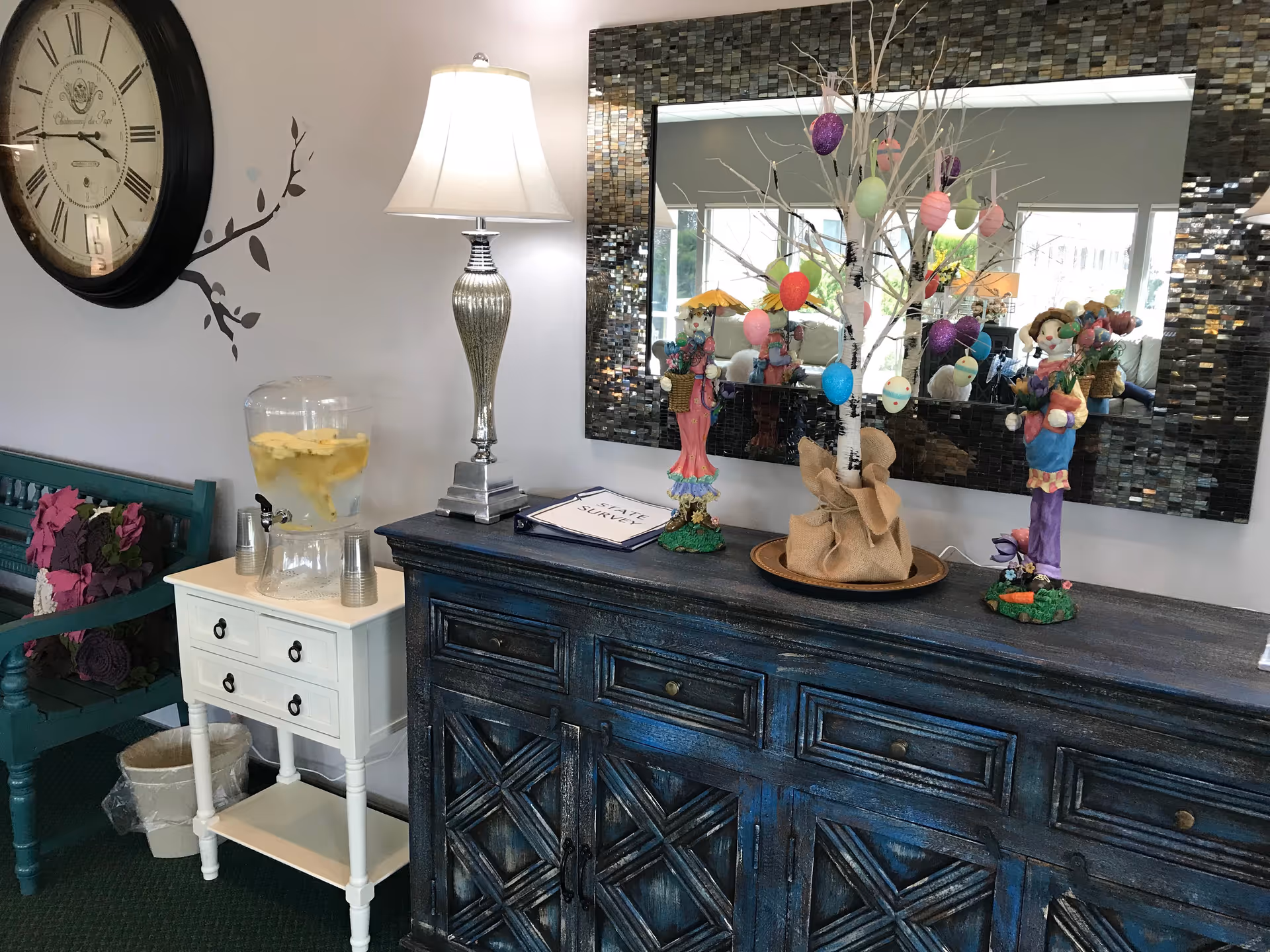 Interior view of a decorated corner in an assisted living facility featuring a large wall clock, a silver table lamp, a water dispenser with cups on a small white table, a dark blue wooden cabinet with decorative figurines and a small tree adorned with colorful Easter eggs, and a large rectangular mirror with a mosaic frame on the wall.