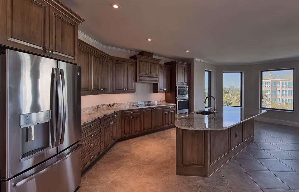 Modern kitchen with dark wooden cabinets, a large island with a granite countertop and sink, stainless steel refrigerator, built-in oven, and large windows showing an exterior view.