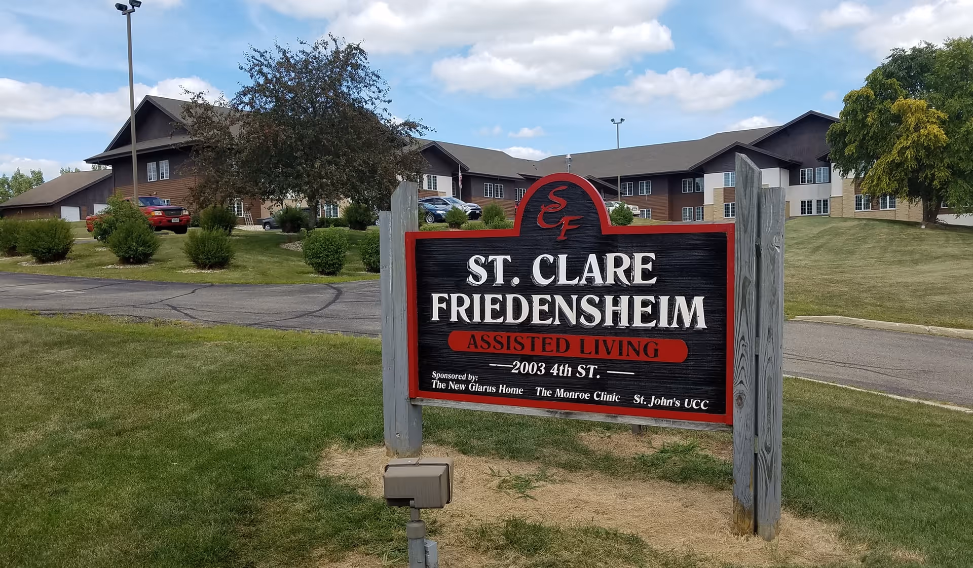 A wooden sign reading 'St. Clare Friedensheim Assisted Living' on a grassy lawn with the assisted living building visible behind it.