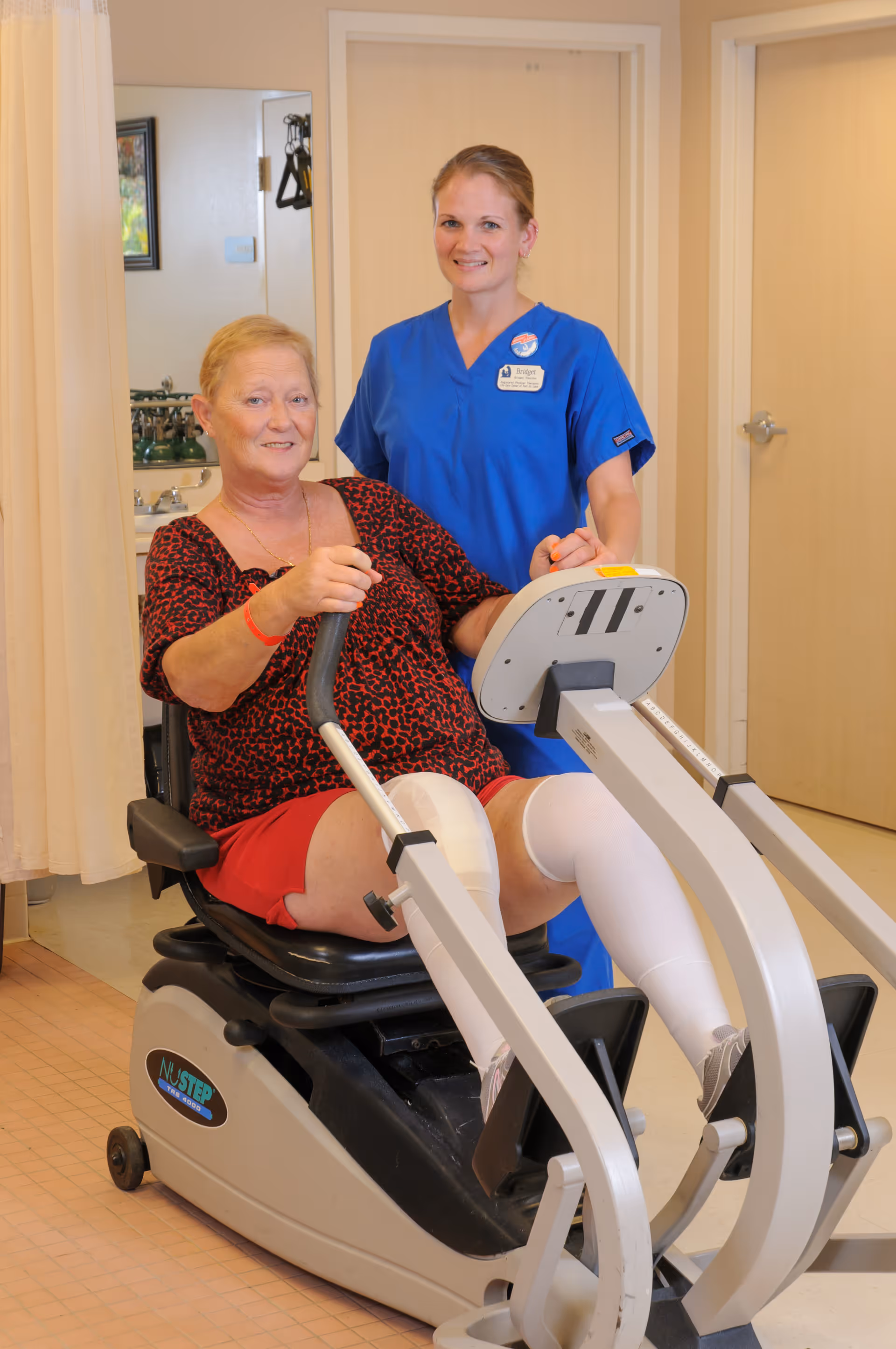 An elderly woman with a prosthetic leg is seated on a NuStep exercise machine, holding the handles. A female healthcare worker in blue scrubs stands beside her, smiling and providing support. The setting appears to be a rehabilitation or therapy room with beige walls and a door in the background.