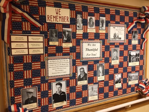 A patriotic bulletin board decorated with small American flags and red, white, and blue ribbons. The board features black and white photographs of veterans along with their names and a message that reads 'We Remember' and 'We Are Thankful For You!'