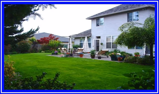 A spacious backyard of a two-story house with a well-maintained green lawn, patio area with outdoor furniture including chairs and umbrellas, surrounded by various plants and trees.