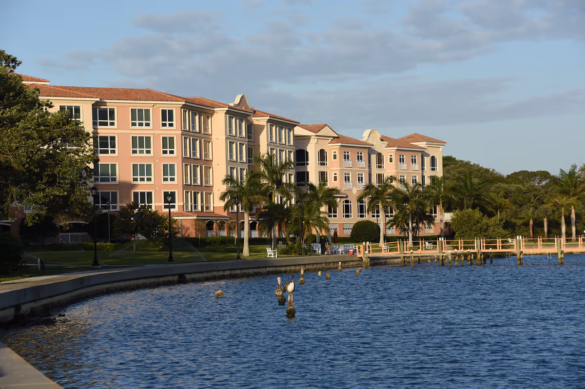 View of a large multi-story residential building with a pink and beige facade, surrounded by palm trees and greenery, situated next to a body of water with a curved concrete walkway along the shore and wooden posts in the water.