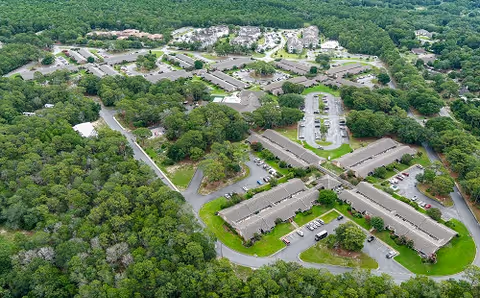 Aerial view of a residential campus with multiple low-rise buildings, parking lots, roads, and surrounding wooded areas.
