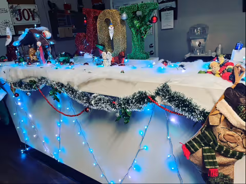 Decorated reception counter with Christmas lights, garlands, 'JOY' letters and holiday figurines in an interior setting.