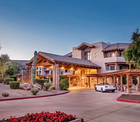 Exterior view of a senior living facility named Vi at Grayhawk during dusk, featuring a covered entrance with stone pillars, a driveway with a white convertible car parked, desert landscaping with cacti and bushes, and a multi-story building with balconies and tiled roofs.