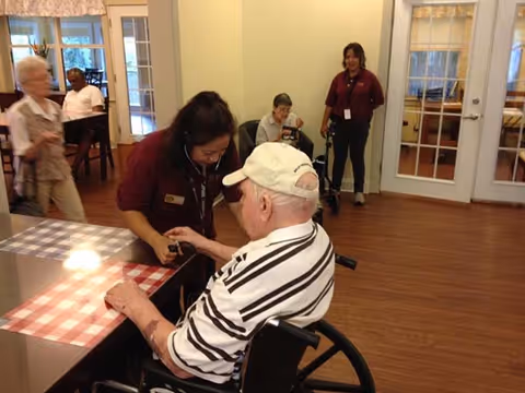 A caregiver in a maroon uniform is interacting with an elderly man in a wheelchair at a table with a checkered tablecloth in a senior living facility. Other elderly residents and staff members are visible in the background in a well-lit room with wooden flooring and glass-paneled doors.