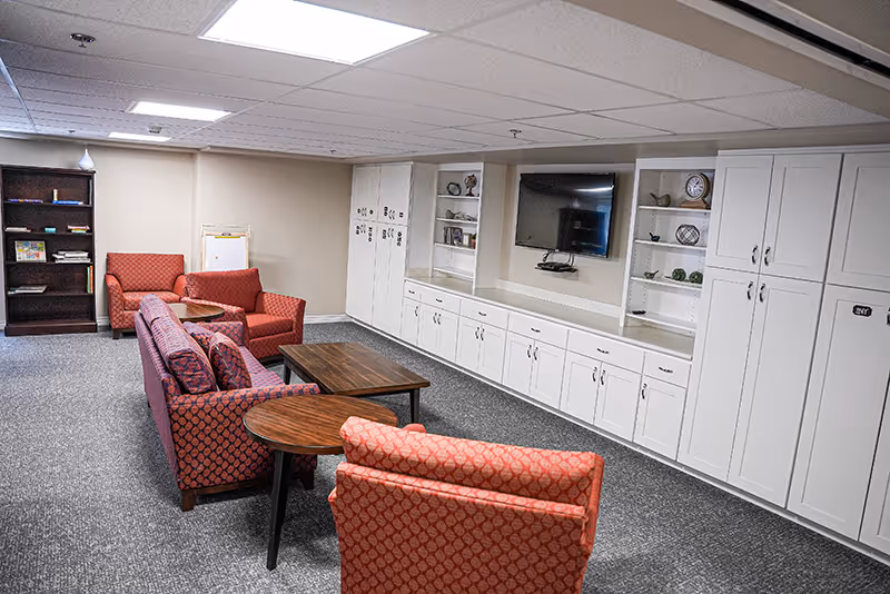 Communal lounge with red upholstered chairs and sofas facing a wall-mounted TV and built-in white cabinetry.