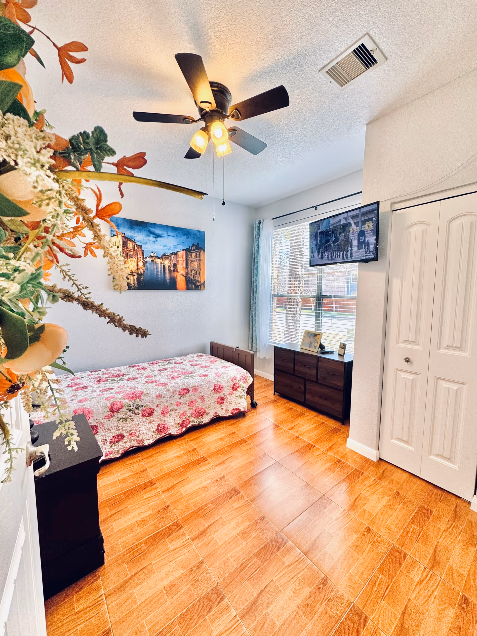 A bright and clean bedroom with a single bed covered in a floral quilt, a ceiling fan with lights, a wall-mounted TV, a window with blinds and curtains, a dark wooden dresser with framed photos, and a closet with white double doors. There is a colorful floral arrangement partially visible on the left side.