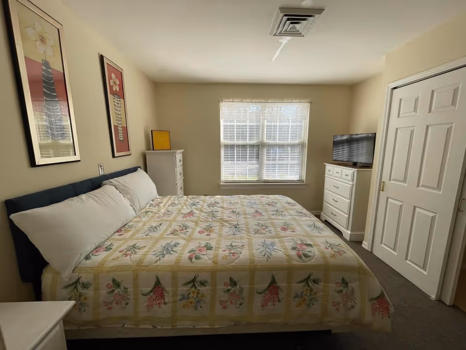 A bedroom with a large bed covered in a floral patterned quilt. There are two framed floral artworks on the wall above the bed. A window with white blinds and lace curtains lets in natural light. A white dresser with a flat-screen TV on top is positioned near a closed closet with sliding doors.