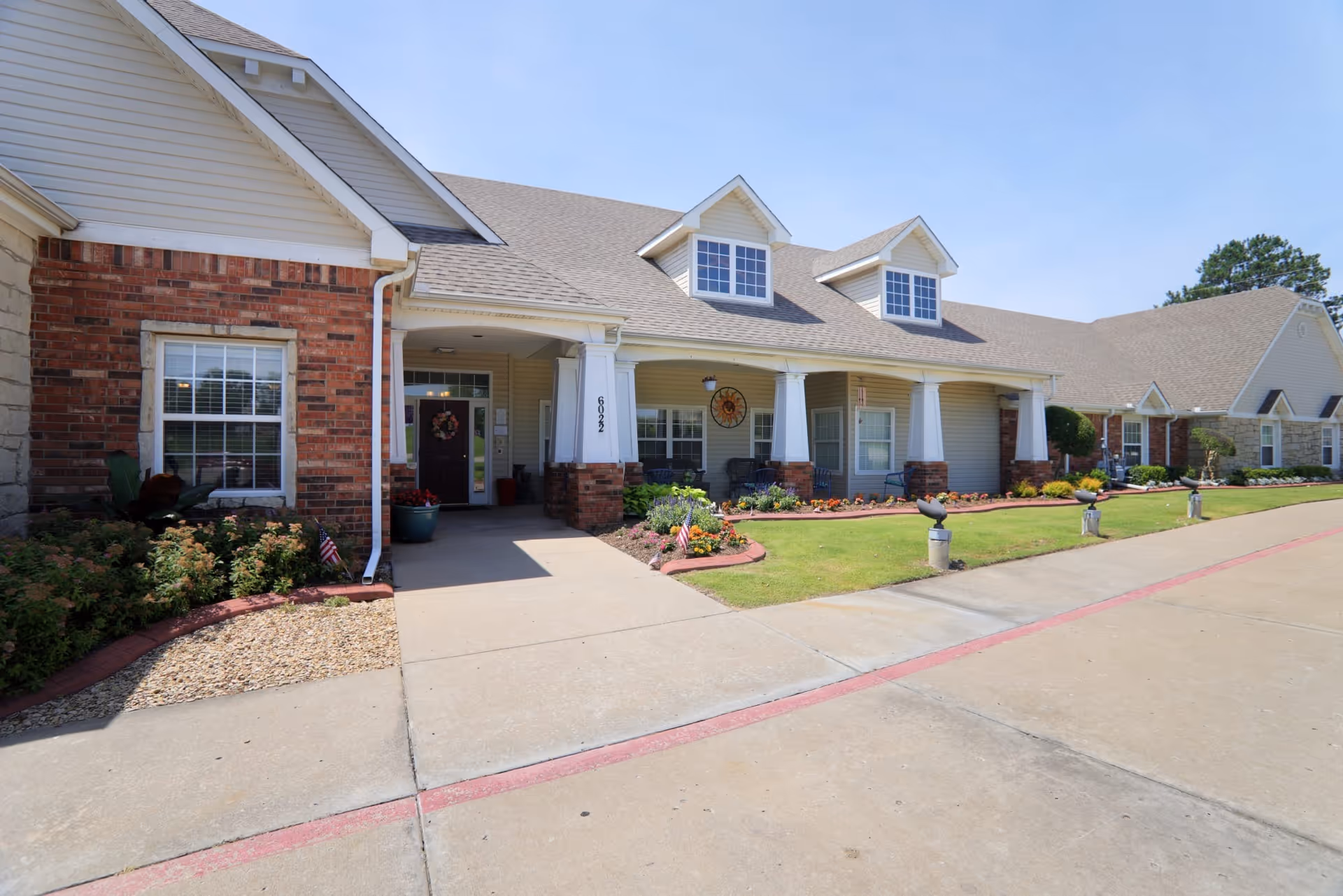 Front entrance of a single-story brick and siding senior living building with a covered porch, landscaped flower beds, and a driveway.