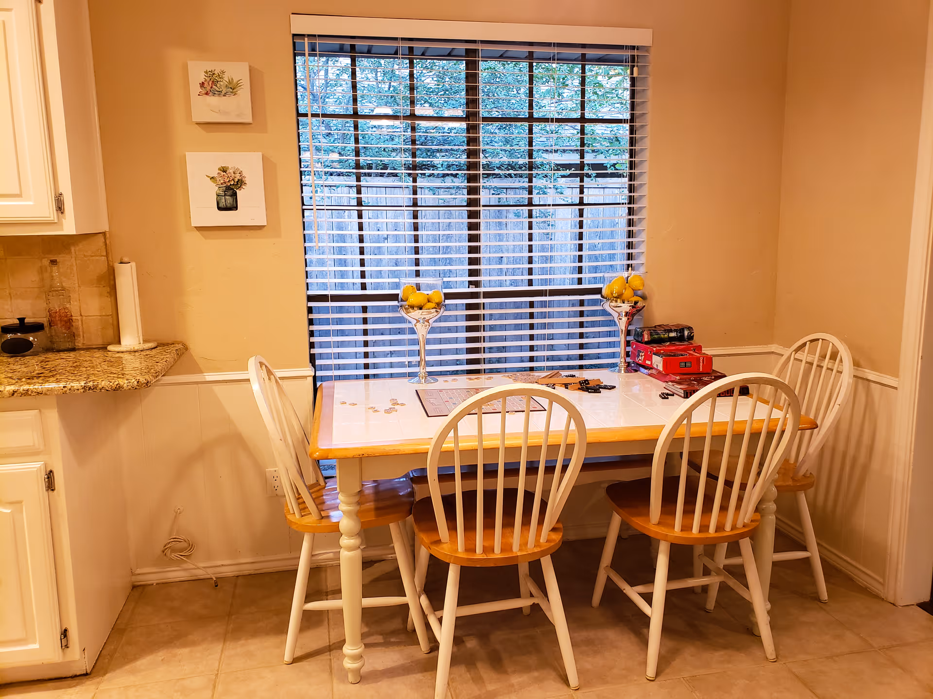 Dining nook with a wooden table and four chairs in front of a large window with blinds, decorated with fruit bowls and small wall art.