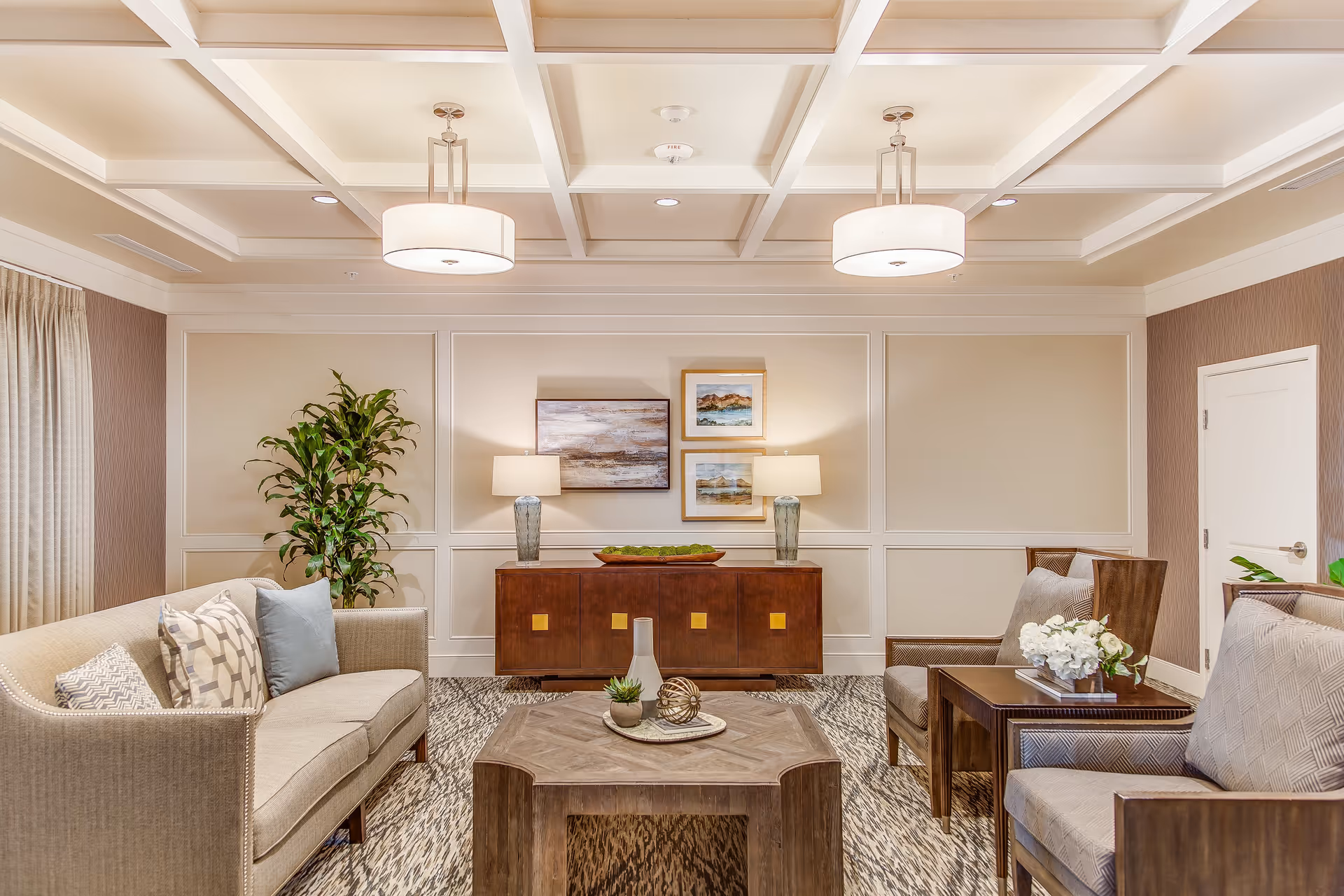 A well-lit living room with a beige sofa and two armchairs arranged around a wooden coffee table. The room features a coffered ceiling with two hanging light fixtures, a sideboard with two table lamps and decorative artwork on the wall above it, and a tall green plant in the corner. The floor is carpeted with a patterned rug.