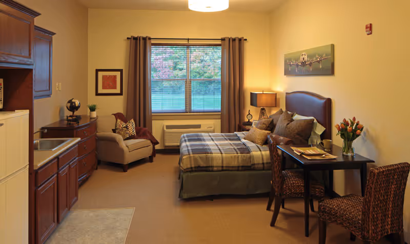 A cozy senior living facility room featuring a bed with plaid bedding, a small dining table with two chairs, a comfortable armchair, a window with brown curtains, and a kitchenette area with a sink and cabinets. The room is warmly lit with a table lamp and decorated with a painting of an airplane and a vase of flowers on the table.