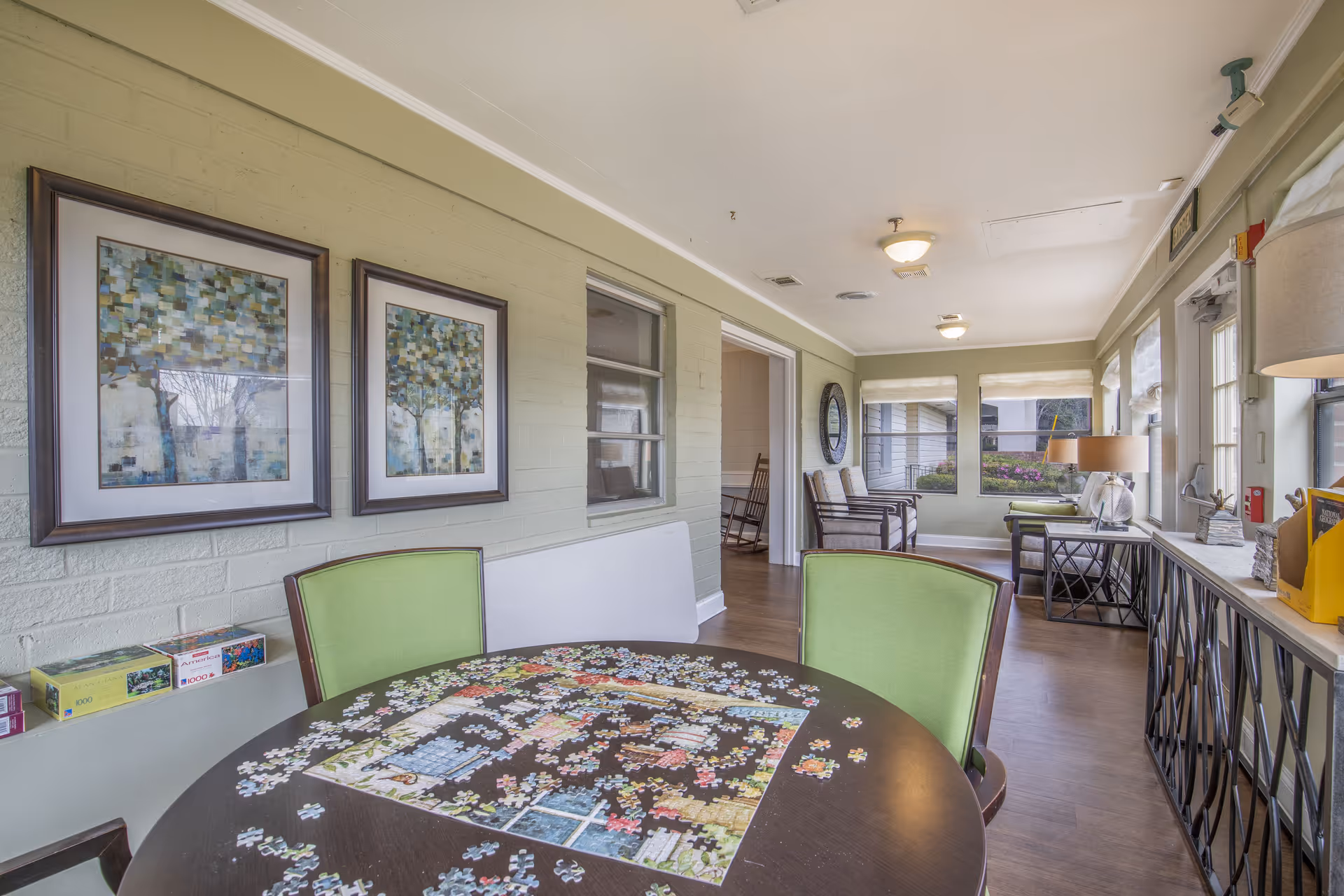 Bright communal lounge in a senior living facility with a round table covered in an unfinished jigsaw puzzle, green chairs, framed artwork, and seating by windows.