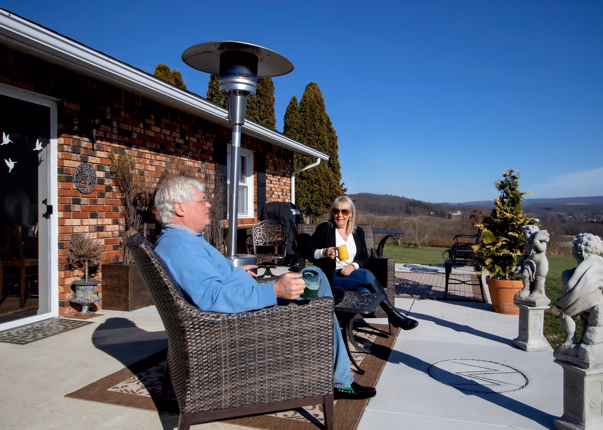 Two elderly people sitting on wicker chairs on a patio outside a brick building, enjoying drinks and conversation on a sunny day with clear blue sky and scenic hills in the background.