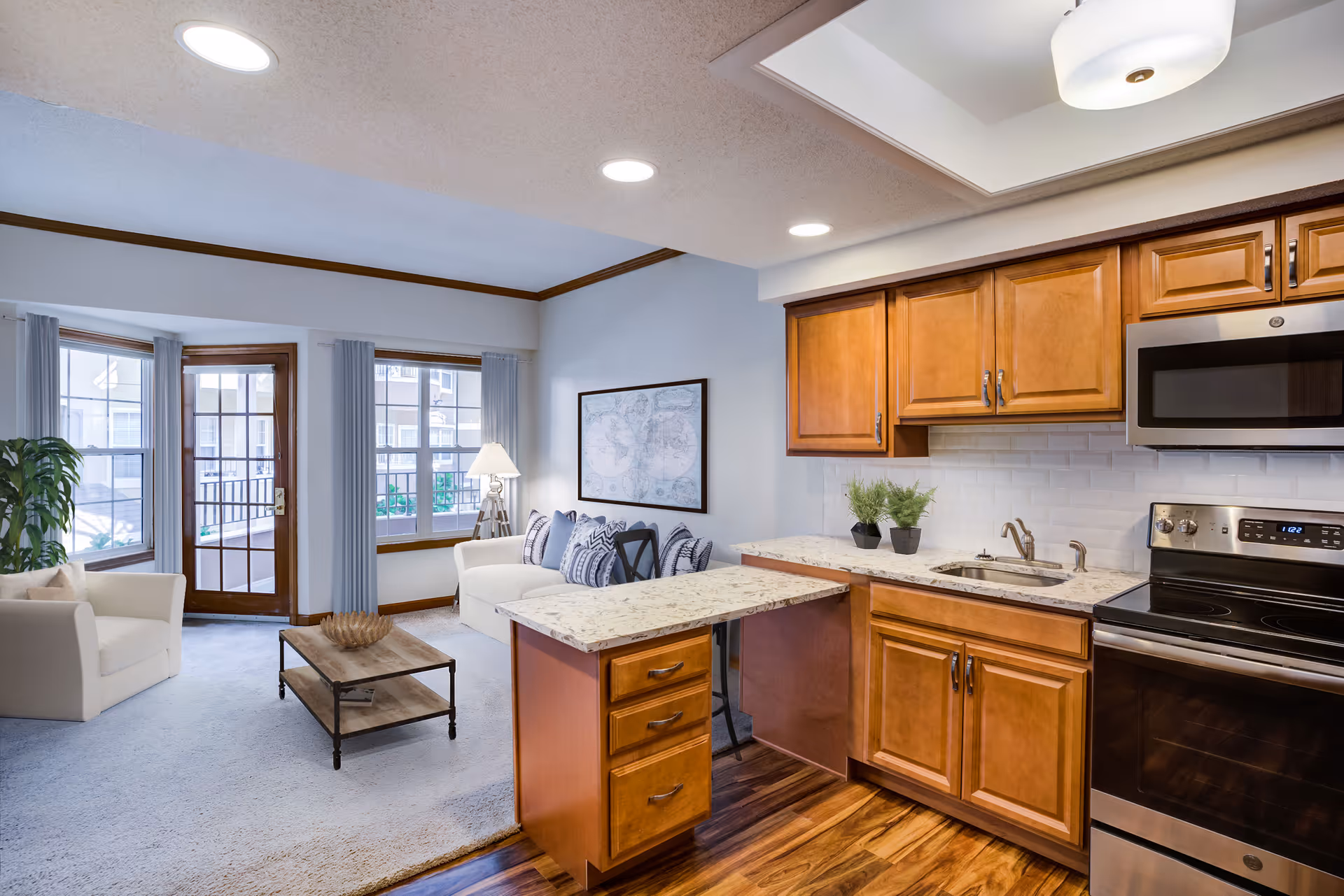 A bright and cozy living area with a small kitchen. The kitchen features wooden cabinets, a stainless steel microwave, stove, and a sink with two small potted plants on the counter. The living area has a white sofa with patterned pillows, a white armchair, a wooden coffee table, a floor lamp, and large windows with vertical blinds letting in natural light. There is a door leading outside and the floor transitions from wood in the kitchen to carpet in the living area.