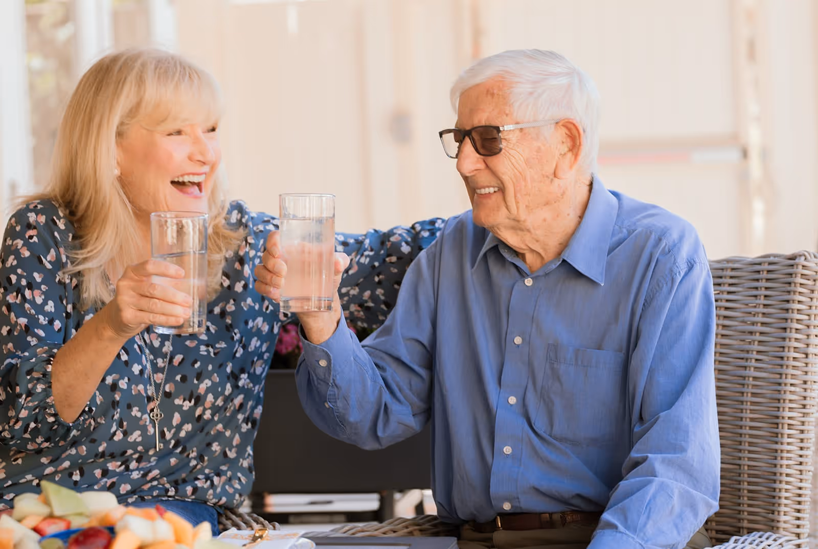 An elderly man and woman sitting outdoors on wicker chairs, smiling and holding glasses of water while toasting each other. There is a plate of cut fruit on the table in front of them.