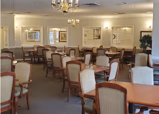 A spacious dining room with multiple wooden tables and cushioned chairs arranged neatly. The room features chandeliers hanging from the ceiling, framed artwork on the walls, and soft lighting from wall sconces. There is a potted plant in the corner and large windows allowing natural light to enter.