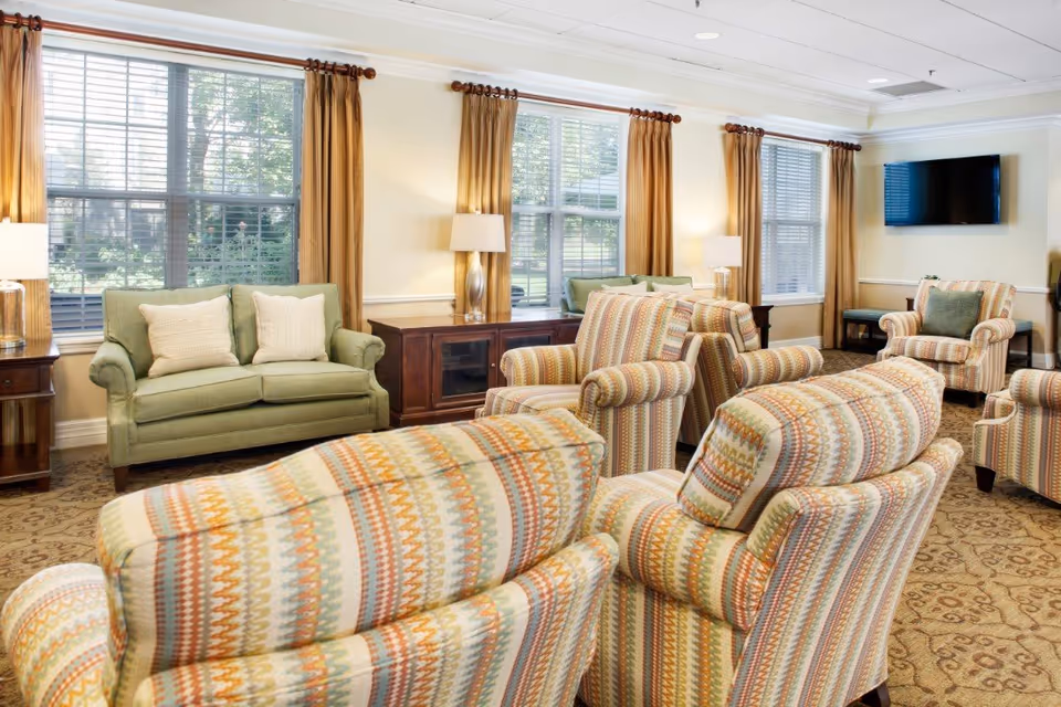 Sunlit communal lounge with patterned upholstered armchairs and a green loveseat arranged around side tables and a wall-mounted TV.