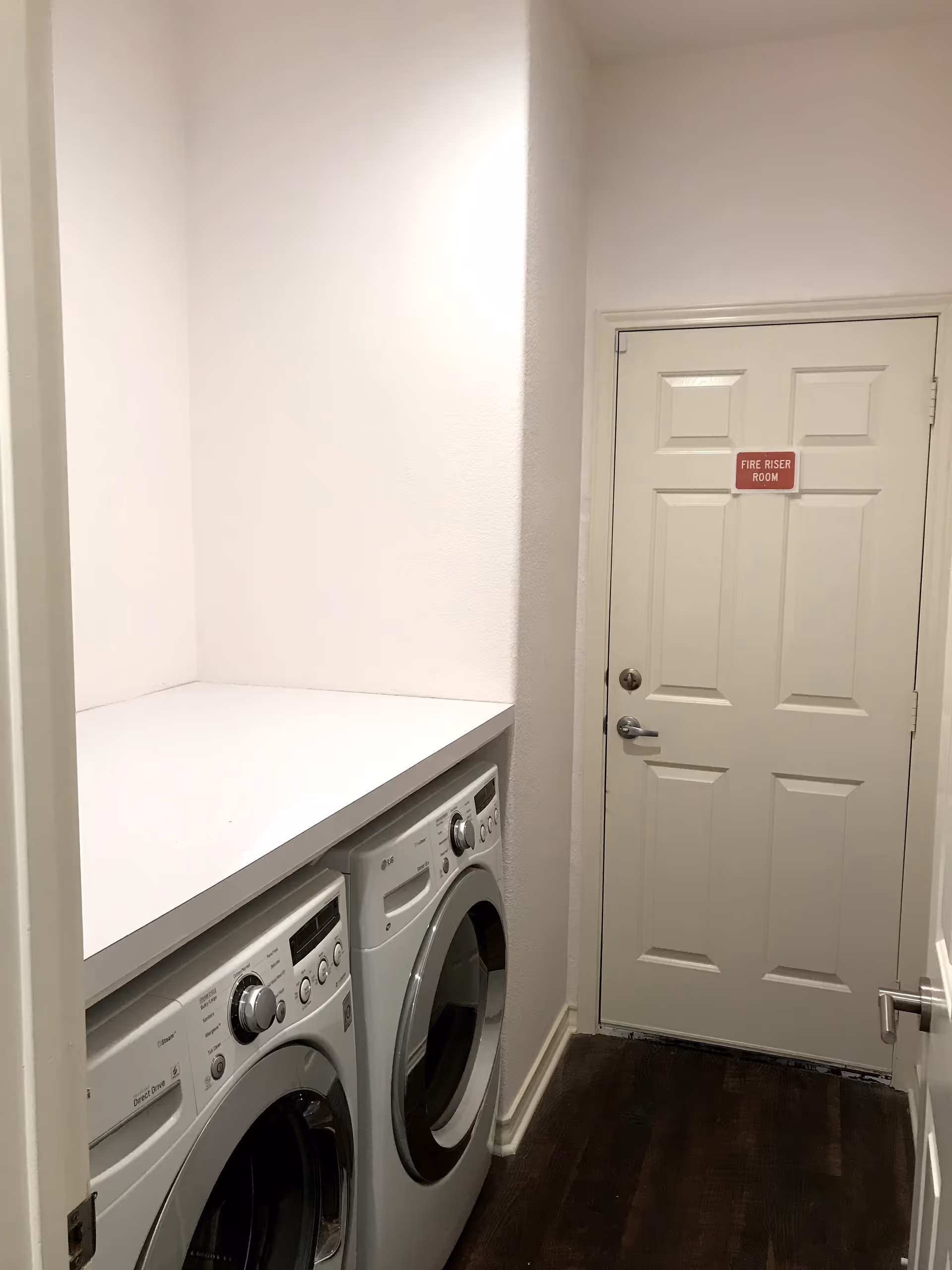 A small laundry room with a white countertop above a front-loading washing machine and dryer. The room has white walls and a door at the end labeled 'FIRE RISER ROOM'. The floor is dark wood.