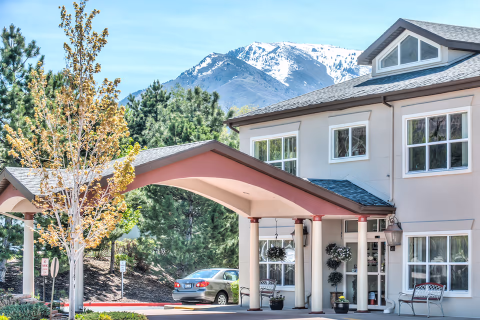 Exterior view of Avamere at Mountain Ridge facility entrance with a covered drop-off area supported by columns, a parked car, benches, potted plants, trees, and snow-capped mountains in the background under a clear sky.