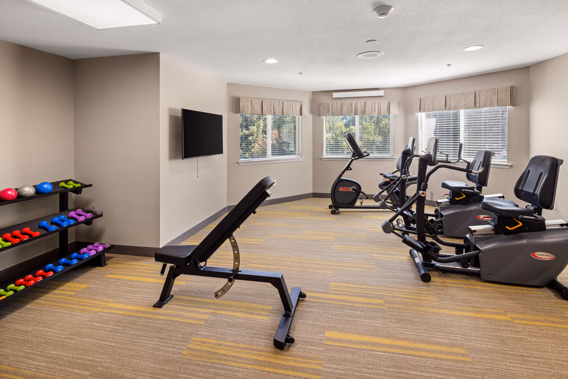 A well-lit exercise room with beige walls and carpeted floor featuring yellow stripes. The room contains a black adjustable workout bench, a rack with colorful dumbbells and exercise balls, and several cardio machines including recumbent bikes and an upright bike. Three windows with beige valances allow natural light to enter the room. A flat-screen TV is mounted on one wall.
