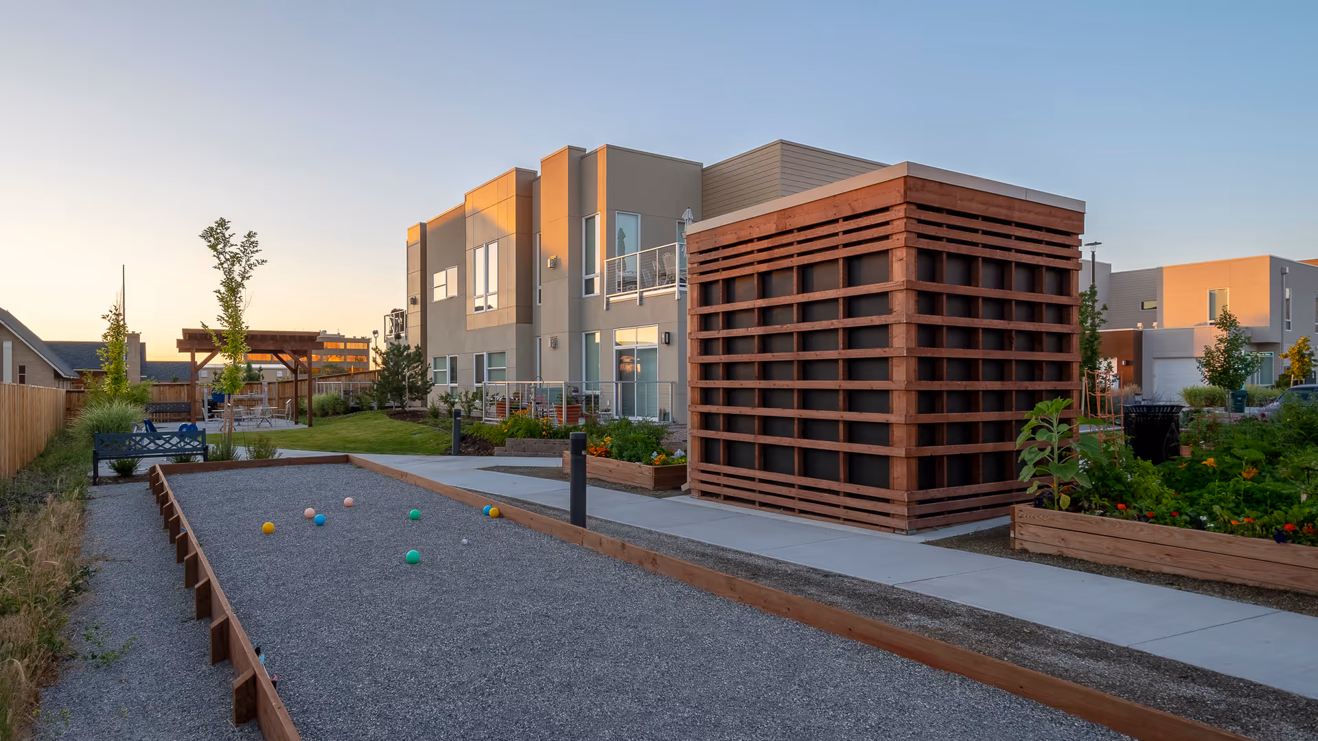 Outdoor area at Village at Belmar featuring a bocce ball court with colorful balls, a wooden pergola with seating, raised garden beds with plants, and a modern two-story building in the background during sunset.
