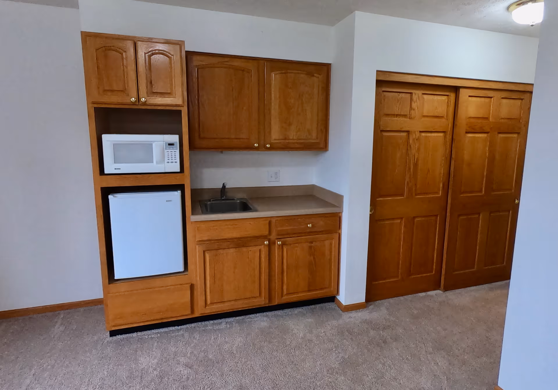 Small kitchenette area with wooden cabinets, a white microwave, a mini refrigerator, a small sink, and a countertop. To the right, there are two wooden sliding closet doors. The floor is carpeted and the walls are painted white.