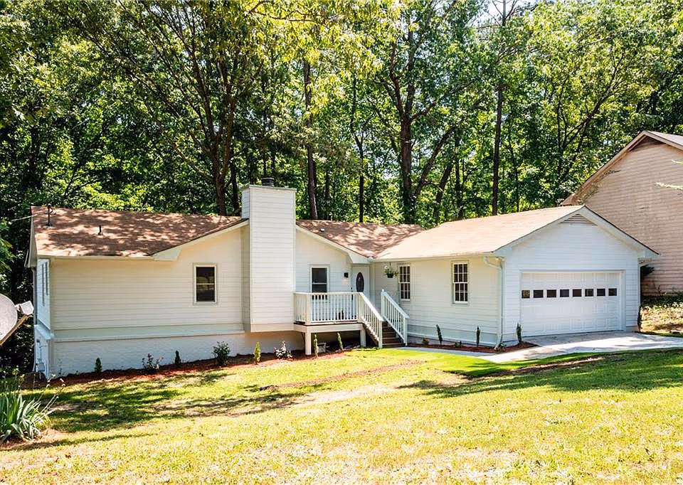 Single-story white house with an attached two-car garage, small front porch and chimney set against a wooded backdrop.