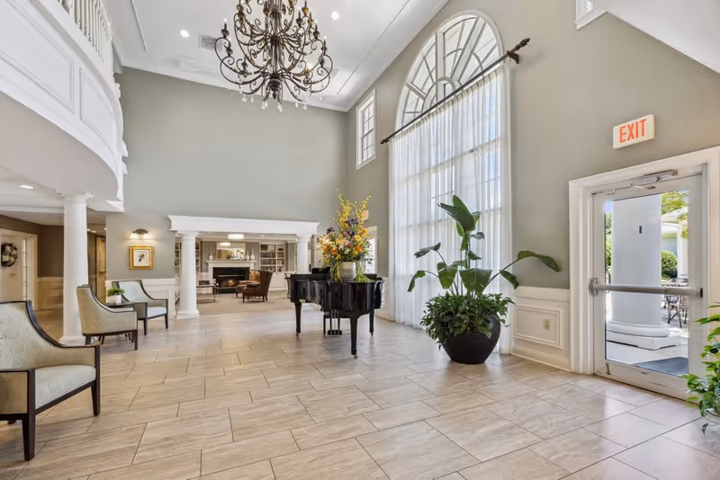 Spacious and elegant senior living facility common area with high ceilings, a large chandelier, a grand piano adorned with a flower arrangement, several chairs along the wall, large arched windows with sheer curtains, and a glass exit door leading outside.