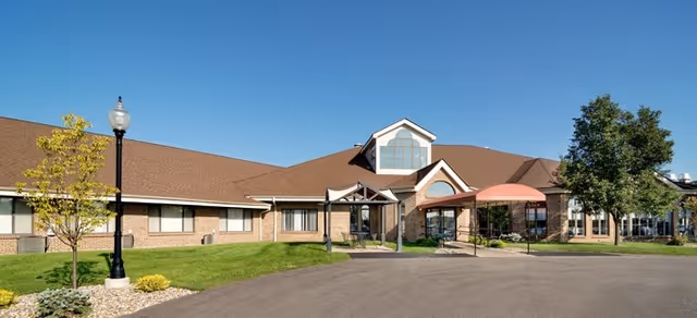 Exterior view of a single-story senior living facility building with a brown roof, brick walls, large windows, a covered entrance with a red canopy, a lamppost, and green landscaping under a clear blue sky.