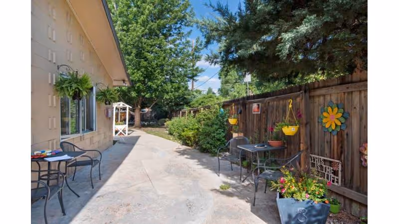 Outdoor patio area with metal chairs and tables along a concrete pathway next to a building. The area is decorated with hanging plants, potted flowers, and colorful wall decorations on a wooden fence. Trees and shrubs are visible in the background under a clear blue sky.