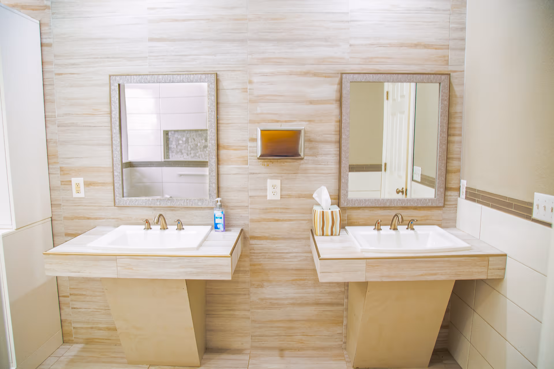 A light-toned double-sink bathroom vanity with two mirrors, a soap dispenser, and a tissue box against a tiled wall.