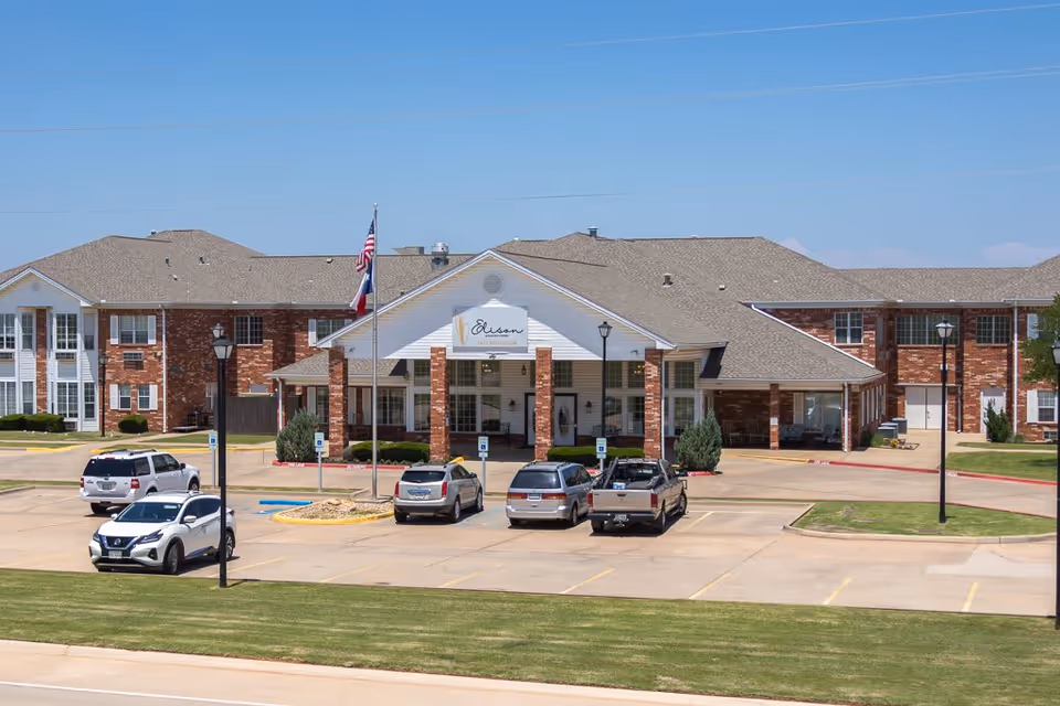 Front exterior view of Elison Assisted Living of Lake Wellington, a large brick building with a covered entrance, several parked cars in front, and two flagpoles displaying the American and Texas flags.