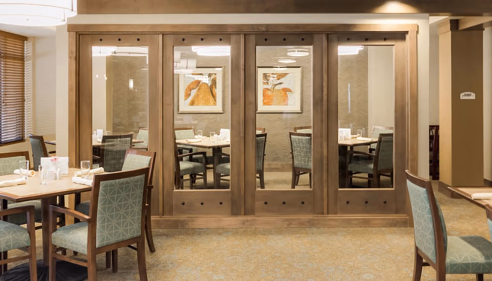 Dining area with wooden tables and chairs upholstered in light blue fabric. The room features large wooden framed glass panels reflecting the interior, with abstract artwork on the far wall. The space is well-lit with natural light coming through window blinds on the left.