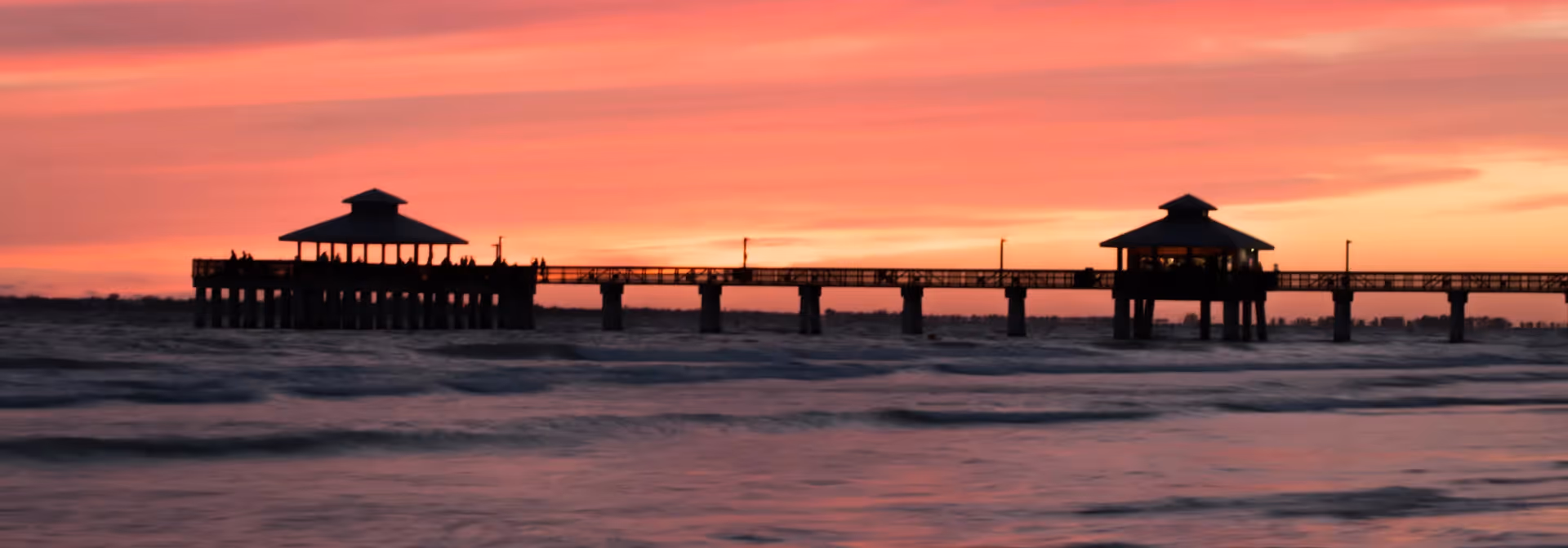 A long pier extending over the ocean with two covered gazebos, silhouetted against a vibrant orange and pink sunset sky.