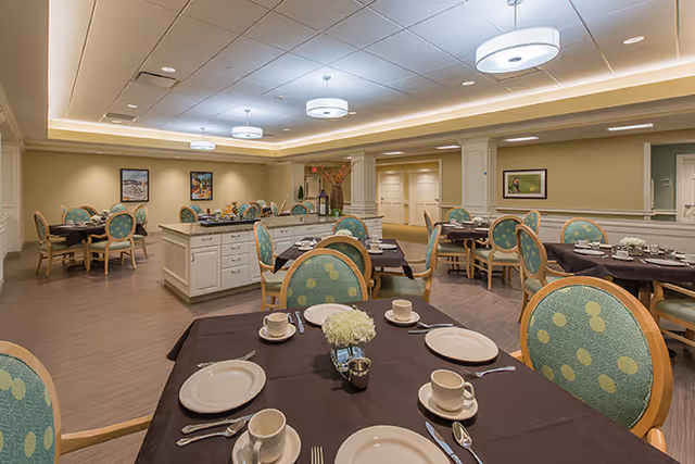 A well-lit dining room in a senior living facility with multiple round tables covered with dark tablecloths, set with white plates, cups, and silverware. The chairs have green upholstery with a circular pattern. The room has beige walls, ceiling lights, and framed artwork on the walls.