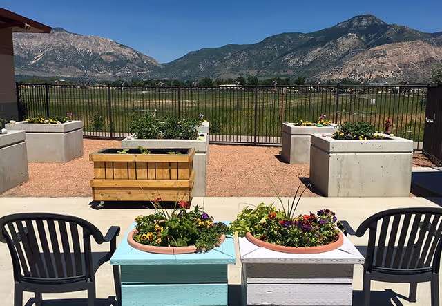 Outdoor patio with chairs and colorful planter boxes overlooking a fenced field and distant mountains.