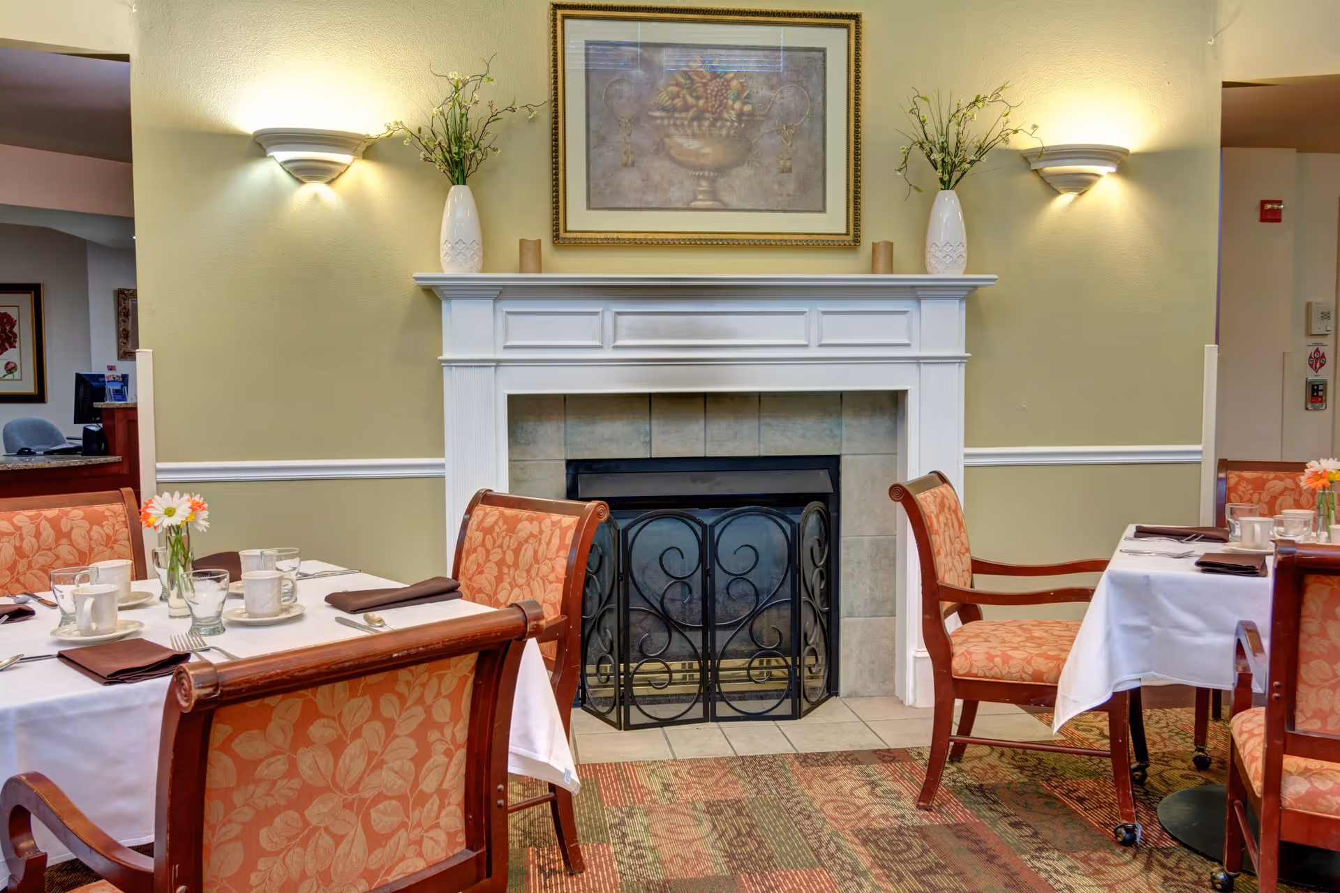 A cozy dining area with tables set with white tablecloths, cups, saucers, and napkins. The room features a white fireplace with a decorative screen, two wall sconces with lights, and a framed painting above the mantel. There are vases with flowers on the mantel and the tables, and upholstered chairs with a floral pattern surround the tables.