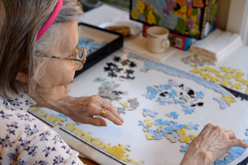 An elderly woman with glasses and a pink headband is working on assembling a jigsaw puzzle on a table. The puzzle pieces are spread out, and the woman is placing pieces on the puzzle board. In the background, there is a coffee cup and the puzzle box.