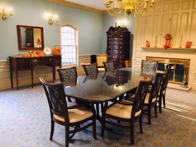 A formal dining room with a large rectangular wooden table surrounded by eight cushioned chairs. The room features light blue walls, a carpeted floor, a chandelier hanging from the ceiling, a fireplace decorated with autumn-themed ornaments, a tall wooden cabinet, and a sideboard with a mirror and seasonal decorations.