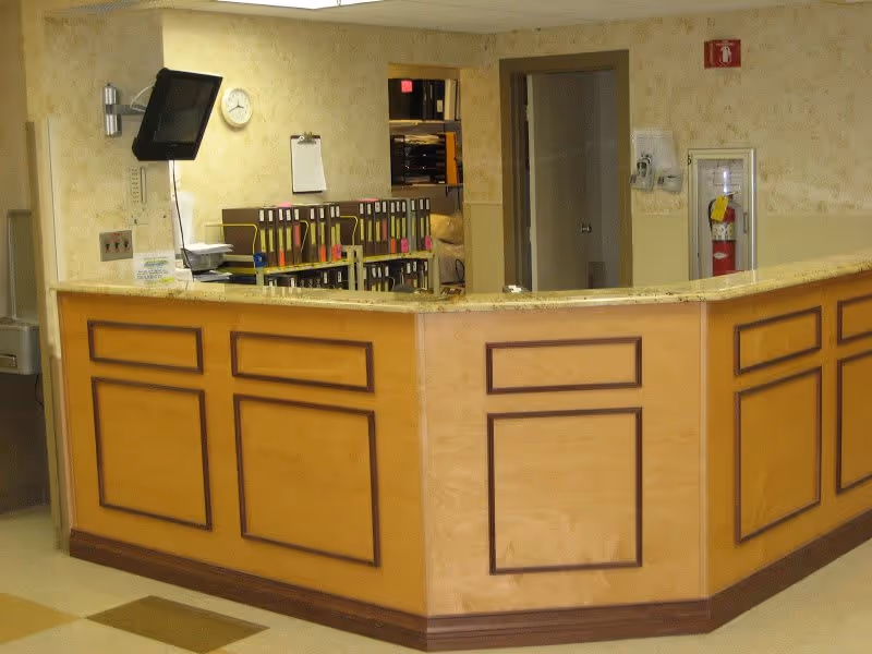 Reception desk area with a wooden counter and beige walls. Behind the desk are shelves with organized files, a wall-mounted clock, a small monitor, and a clipboard. A fire extinguisher is mounted on the wall to the right.