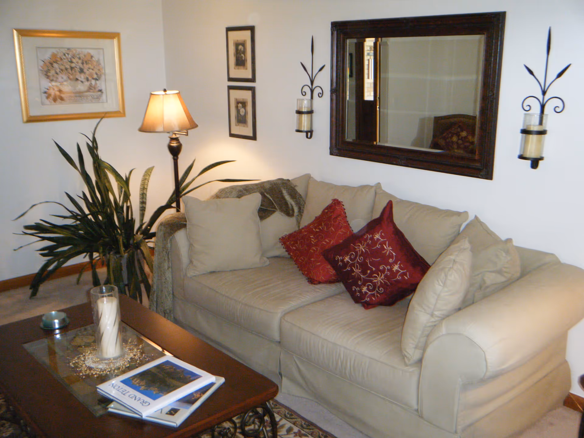 A cozy living room area with a beige sofa adorned with red and beige decorative pillows. A large mirror hangs on the wall above the sofa, flanked by two wall-mounted candle holders. A floor lamp and a large green plant are positioned next to the sofa. In front of the sofa is a wooden coffee table with a glass top, holding a large candle and some books. The walls are decorated with framed artwork.