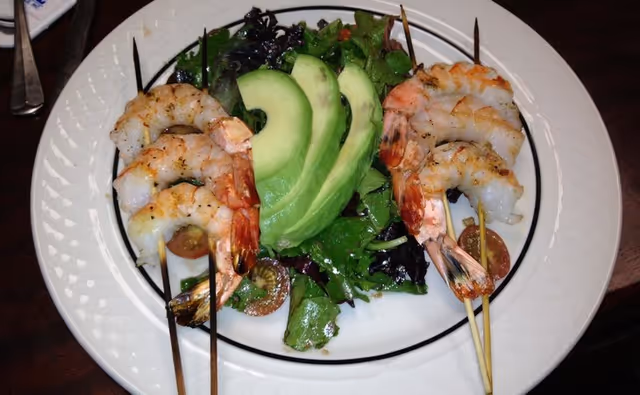Grilled shrimp skewers served over mixed greens with avocado slices and cherry tomatoes on a white plate.