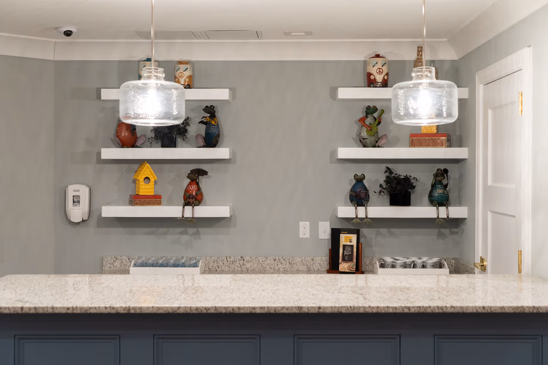 Interior view of a countertop with a granite surface and blue cabinetry below. Above the counter, two pendant lights with glass shades hang from the ceiling. On the wall behind the counter, there are two sets of white floating shelves displaying decorative frog figurines, a yellow birdhouse, small plants, and other decorative items. A white door is visible on the right side of the image, and a hand sanitizer dispenser is mounted on the left wall.