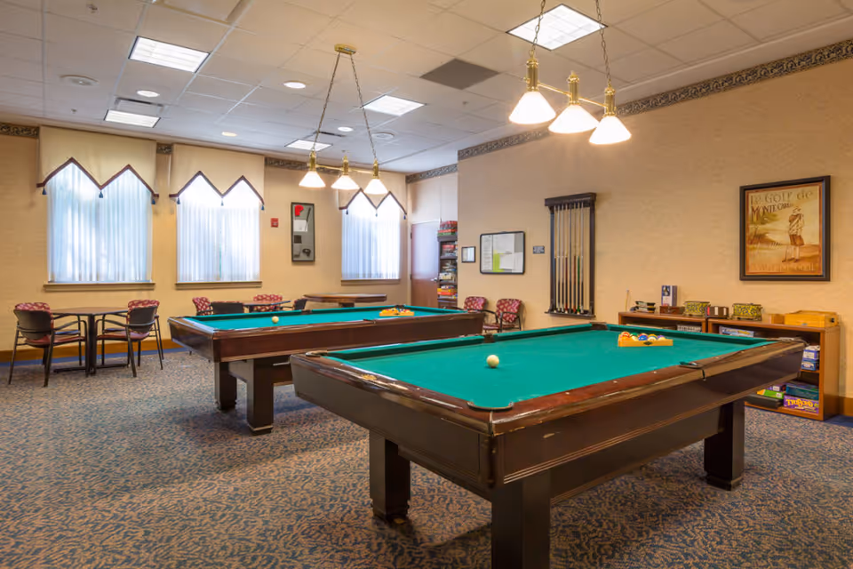 A recreational room with two pool tables, several chairs, and small tables near windows with white curtains. The room has carpeted flooring, beige walls, and ceiling lights. There are pool cues mounted on the wall and a framed poster.
