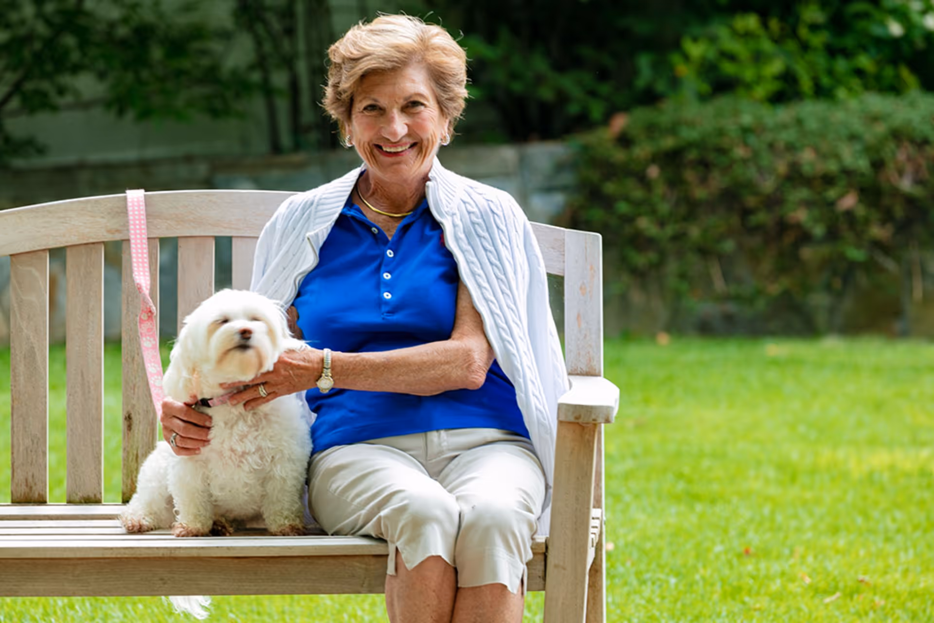 An elderly woman sitting on a wooden bench outdoors in a garden, smiling and holding a small white dog on her lap. She is wearing a blue shirt, white pants, and a white sweater draped over her shoulders. The background shows green grass and bushes.